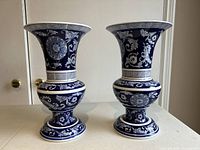 Photo shows two matching blue vases with detailed white floral and Greek key patterns. Each vase has a flared lip and sits on a rounded base.