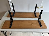 Two wooden shelves placed on a white folding table, showing the black metal bracket support structures attached underneath each shelf.