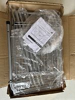 Opened box showing silver metal wire shelving parts, support rods, and instruction manual wrapped in plastic