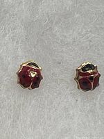 Front view of the pair of 10K gold ladybug stud earrings with red and black enamel on a light fabric background.