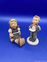 Both Goebel Hummel figurines displayed together against a blue background to show size and styling.