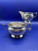 View showing the hand hammered sugar bowl and cream pitcher with distinct round footed bases and handles against blue background.