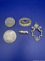 Five vintage silver items arranged on a blue surface including circular Aztec calendar pin, oval Mexican pin with lapis stone, running boar brooch, large ornate brooch, and round brooch with cowboy motif
