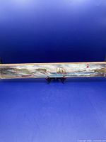Full view of the long narrow seaside wood painting on a display stand with lighthouse, ship, shore and water details.