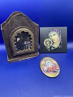 Full view of the three religious items against a blue background: wooden plaque, square tile, oval wall hanging