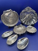 Seven silver plated shell shaped dishes arranged on a blue surface with some standing on small dish holders showing various sizes and shapes.