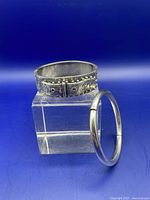 Both bracelets shown together; one is a plain sterling bangle, the other a buckle-style sterling bracelet with decorative texture.