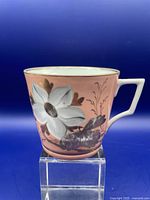 Side view of vintage moustache cup showing pink color with white flower and gilt accents, square handle.