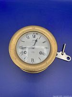 Front view of round brass ship's bell clock with dial showing Roman numerals and winding key beside it.