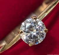 Close-up front view of round moissanite stone set with four prongs in 10K yellow gold band on a red background.