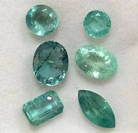 Six loose emerald gemstones in various cuts on a white background showing natural inclusions and green color variety.