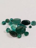 Close-up photo showing multiple loose emerald gemstones in a variety of green shades, cuts, and sizes on a white surface.