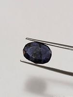 Oval faceted genuine iolite gemstone held with tweezers, showing dark blue-violet color and natural inclusions.