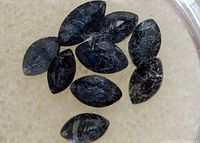 Top down view of eight marquise-shaped dark blue sapphire gemstones with visible inclusions on a light background.