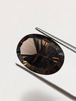 Close-up image of an oval faceted smokey quartz gemstone held by tweezers against a white background, showing its deep smoky brown color and faceting details.
