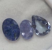 Three tanzanite gemstones including one pear-shaped faceted stone and two oval stones with varying transparency and color intensity, arranged on a light surface.