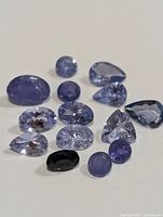 Photo of 14 loose tanzanite gemstones laid out on a white surface showing various shapes and colors predominantly bluish-violet.