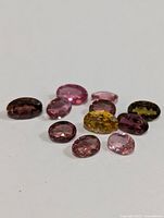 Ten faceted oval-shaped tourmaline gemstones in varying colors including pink, yellow, dark green, and brown laid on a white background.