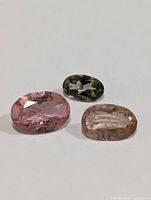 Three oval faceted tourmaline gemstones in pink, green, and peach shown on white background