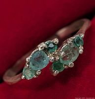 Close-up angled view of the ring showing the emerald stones cluster and silver setting on red velvet background.
