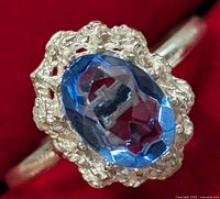 Close-up of the silver ring featuring an oval faceted blue gemstone in a textured decorative bezel setting on a plain band.