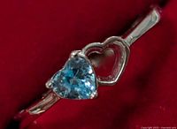 Close-up photo of a sterling silver ring with a heart-shaped blue gemstone and a hollow heart accent on the band, displayed on red fabric.