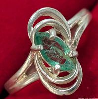 Close-up front view of silver ring with marquise emerald set in artistic swirl silver band on red background.