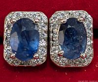 Close-up front view of silver stud earrings with oval blue sapphires and diamond halo on red background.