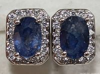 Front view of the pair of silver earrings with oval blue sapphires and clear stones around them.