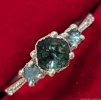 Close-up of the ring showing the center dark green stone and two smaller blue stones on either side, set in a silver band.