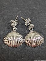 Front view of two silver shell-shaped dangle earrings on a dark background showing detailed ridges and shiny finish.