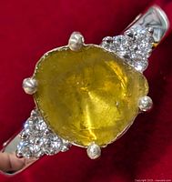Close-up of front of ring showing yellow center stone and clear accent stones on a silver band against a red background.