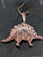 Top and side angled view of silver stegosaurus pendant showing dinosaur shape with detailed back plates and tail, on black background.