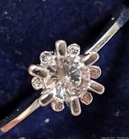 Close-up of sterling silver ring with central clear faceted stone and six prongs, on a blue fabric background.