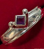 Front view of silver ring with square-cut amethyst set in an angular silver frame with bead accents on a red background.
