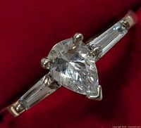 Close-up image of silver ring with pear-shaped clear center stone and baguette side stones on a red background.