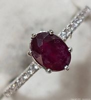 Close-up of ring showing oval deep red ruby center stone with six prong setting and small clear CZ stones along band shoulders.