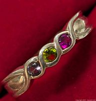 Close-up photo of a sterling silver ring with three bezel-set round stones in purple, green, and pink colors on a twisted band against a red background.