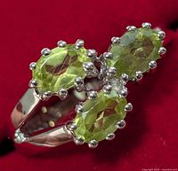 Close-up photo of silver ring with three oval peridot stones set in prong settings against a red background.