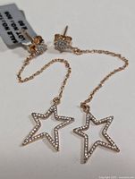 Close-up of both silver earrings showing the star stud with white topaz and the dangling open star pendant with small sparkling stones connected by a fine chain.