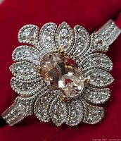 Close-up front view of silver ring showing central oval Morganite CZ stone, surrounded by petal-shaped halo of small clear stones and ring band with small stones and milgrain detail.