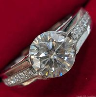 Close-up of silver ring with large round moissanite stone and accent stones on the sides against a red background.