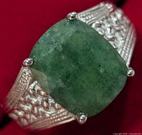 Close-up view of the cushion-cut green emerald and intricate silver band with woven details and four prongs holding the gemstone.