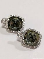 Close-up of front face of the two silver earrings each with a green faceted gemstone and halo of clear stones.
