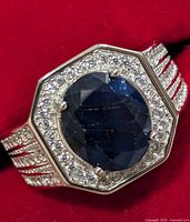 Close-up photo of the silver ring showing the detailed face with oval sapphire and surrounding clear stones on red background.