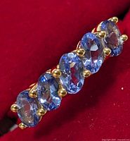 Close-up image showing side angle of silver ring with five oval tanzanite gemstones held by gold-toned prongs on a red background.