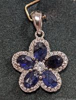 Top view of silver flower pendant showing five oval blue sapphires and central clear stone with surrounding small clear stones on dark fabric background.