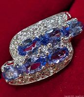 Close-up of the silver ring featuring multiple oval tanzanite stones mounted in an intricate design, showing prong settings and textured silver band on a red background.