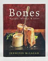 Front cover of 'Bones: Recipes, History & Lore' hardcover cookbook by Jennifer McLagan, showing three cooked marrow bones on a black plate with a spoon.