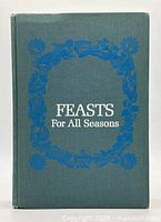 Front cover of the hardcover cookbook 'Feasts For All Seasons' by Roy Andries de Groot with a decorative blue floral wreath on green cloth binding.